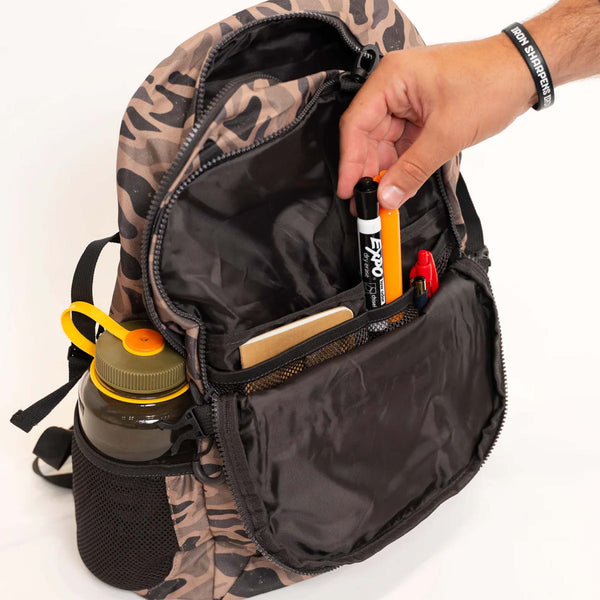 Burlebo Backpack in Guage Camo, open with a hand placing markers in the front organizer pocket. Mesh side pocket holds a water bottle.
