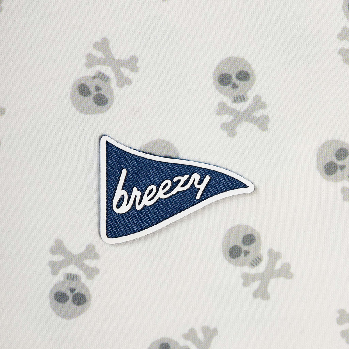 Close-up of Breezy white polo fabric featuring gray skull and crossbones pattern with navy pennant logo patch.