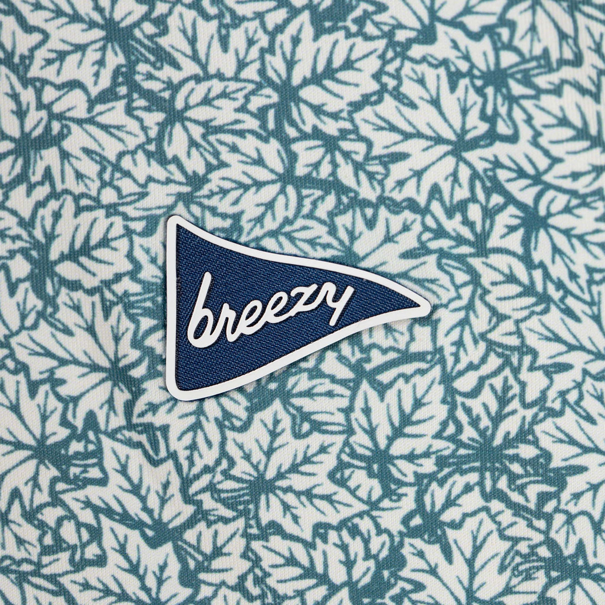 Breezy men’s performance polo in white and green leaf print with close-up of embroidered navy pennant logo on chest.