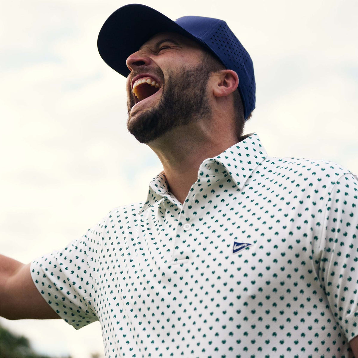 Man wearing a blue cap and white polo with all-over small clover pattern outdoors and laughing