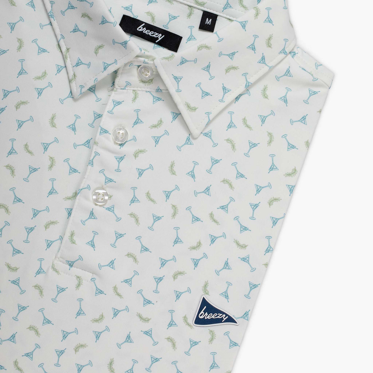 Folded men's white polo with an all over print of small martini glasses in blue and green shown on a white background