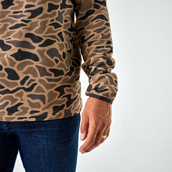 Close-up of men’s brown and black camo quarter-zip hoodie hemline and sleeve, paired with dark blue jeans.