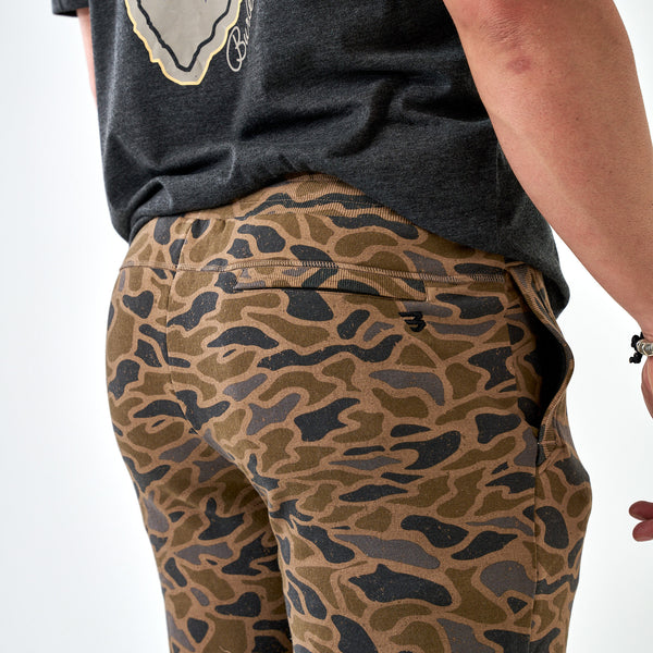 Full-length side angle of men’s camo jogger pants in green and tan camo print, shown with sneakers and dark tee.