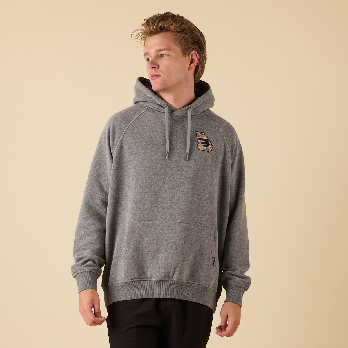 Front view of a man wearing a gray hoodie with a small camo-pattern logo on the left chest, looking to the side.