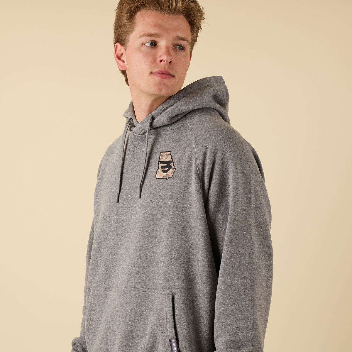 Angled front view of a man in a gray hoodie with a small camo-pattern logo on the left chest, looking away from the camera.