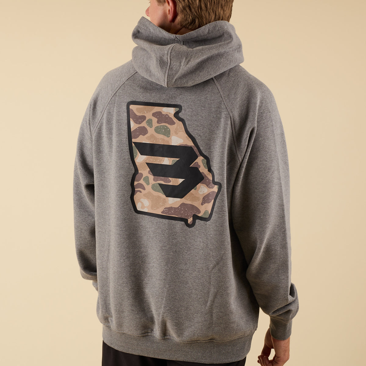 Rear angled view of a man wearing a gray hoodie with a large camo-pattern logo graphic on the back.