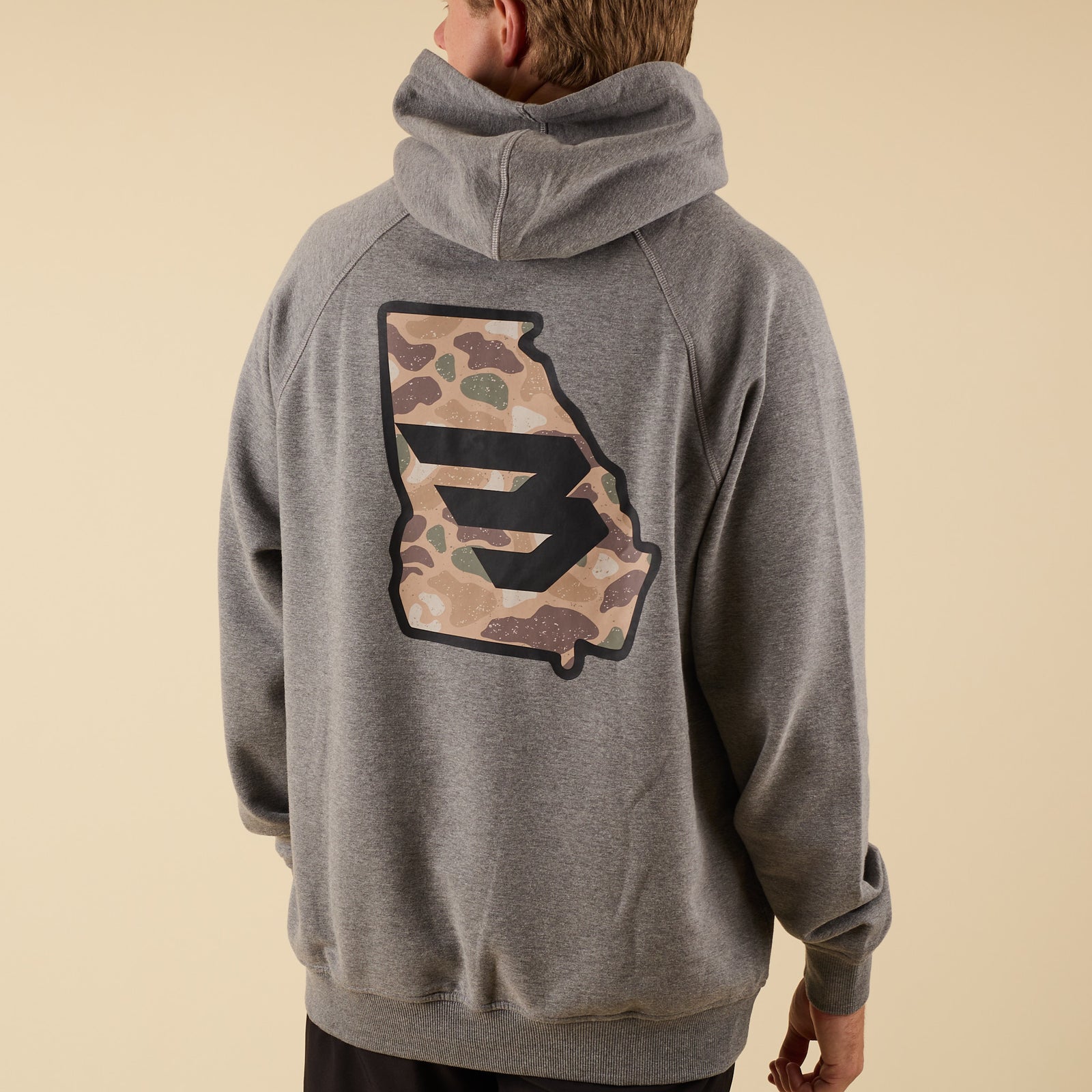 Rear angled view of a man wearing a gray hoodie with a large camo-pattern logo graphic on the back.