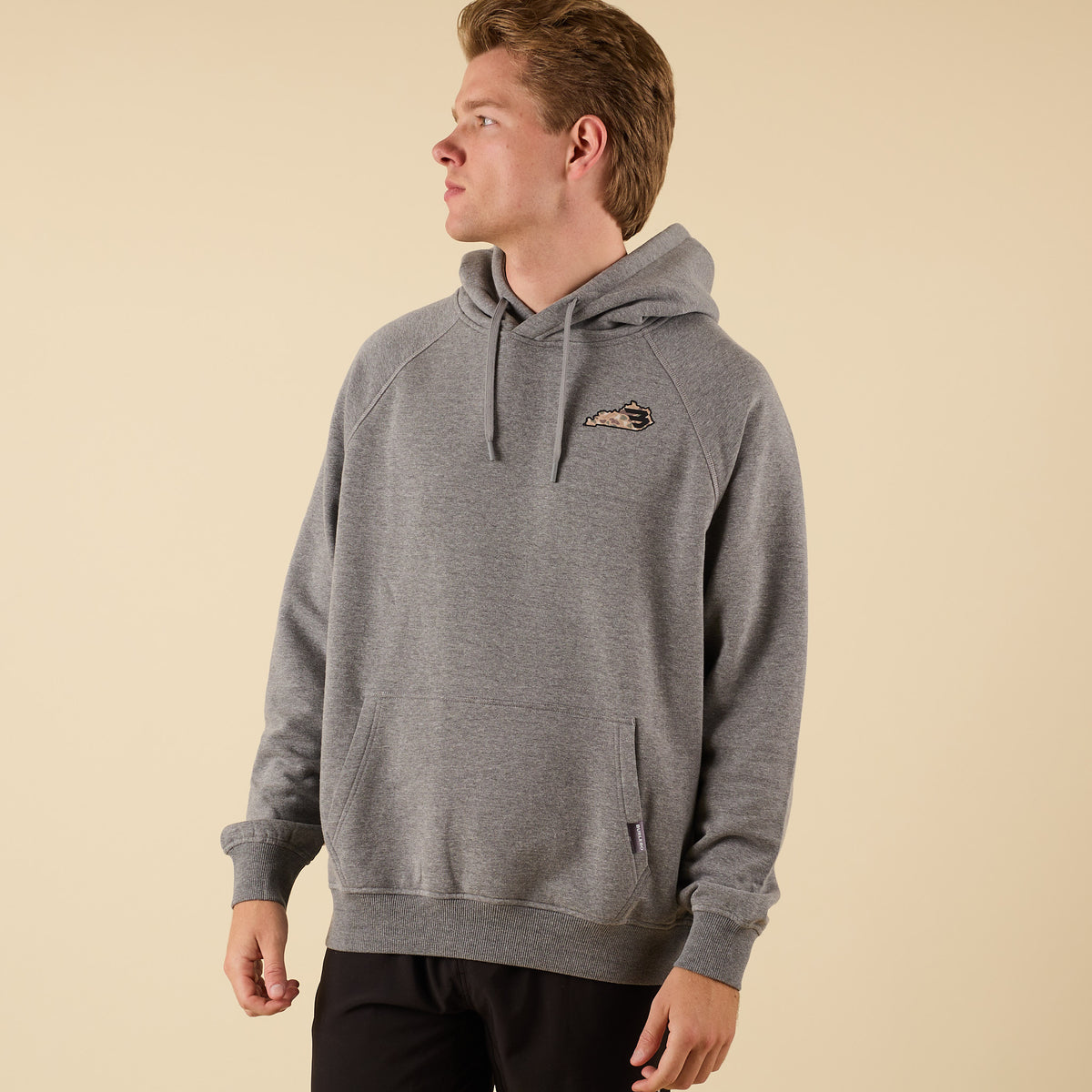 Front view of a man wearing a gray hoodie with a small camo-pattern logo on the left chest, looking to the side.