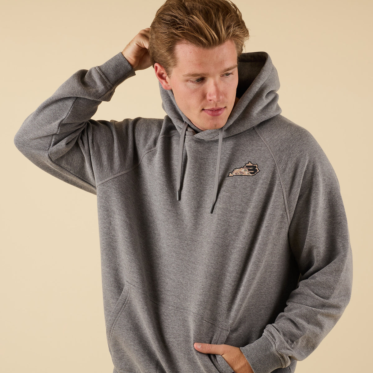 Close-up front view of a man in a gray hoodie with a small camo-pattern logo on the left chest, one hand adjusting his hair and the other in the pocket.