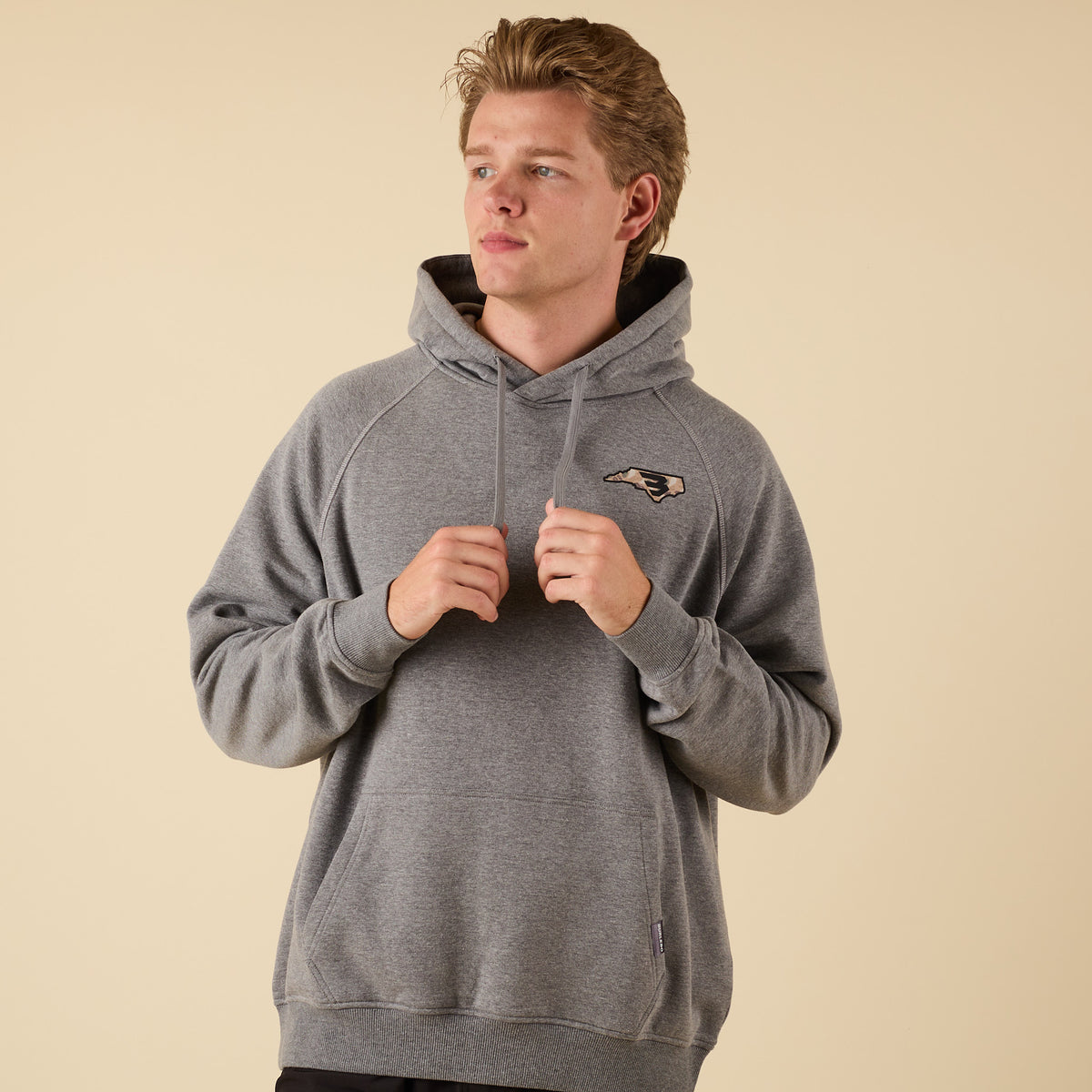 Front view of a man wearing a gray hoodie with a small camo-pattern logo on the left chest, holding the hood drawstrings.