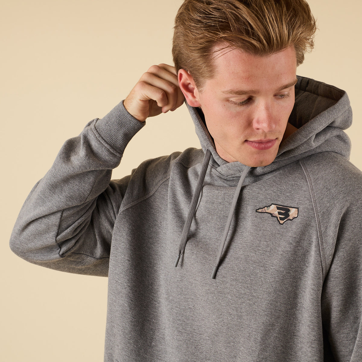 Close-up front view of a man in a gray hoodie with a small camo-pattern logo on the left chest, adjusting the hood.