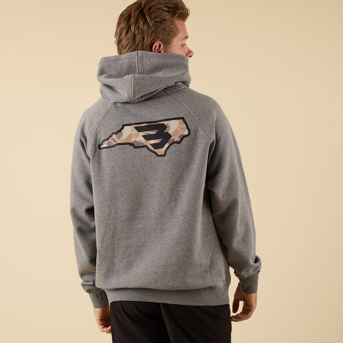 Rear angled view of a man wearing a gray hoodie with a large camo-pattern logo graphic centered between the shoulders.