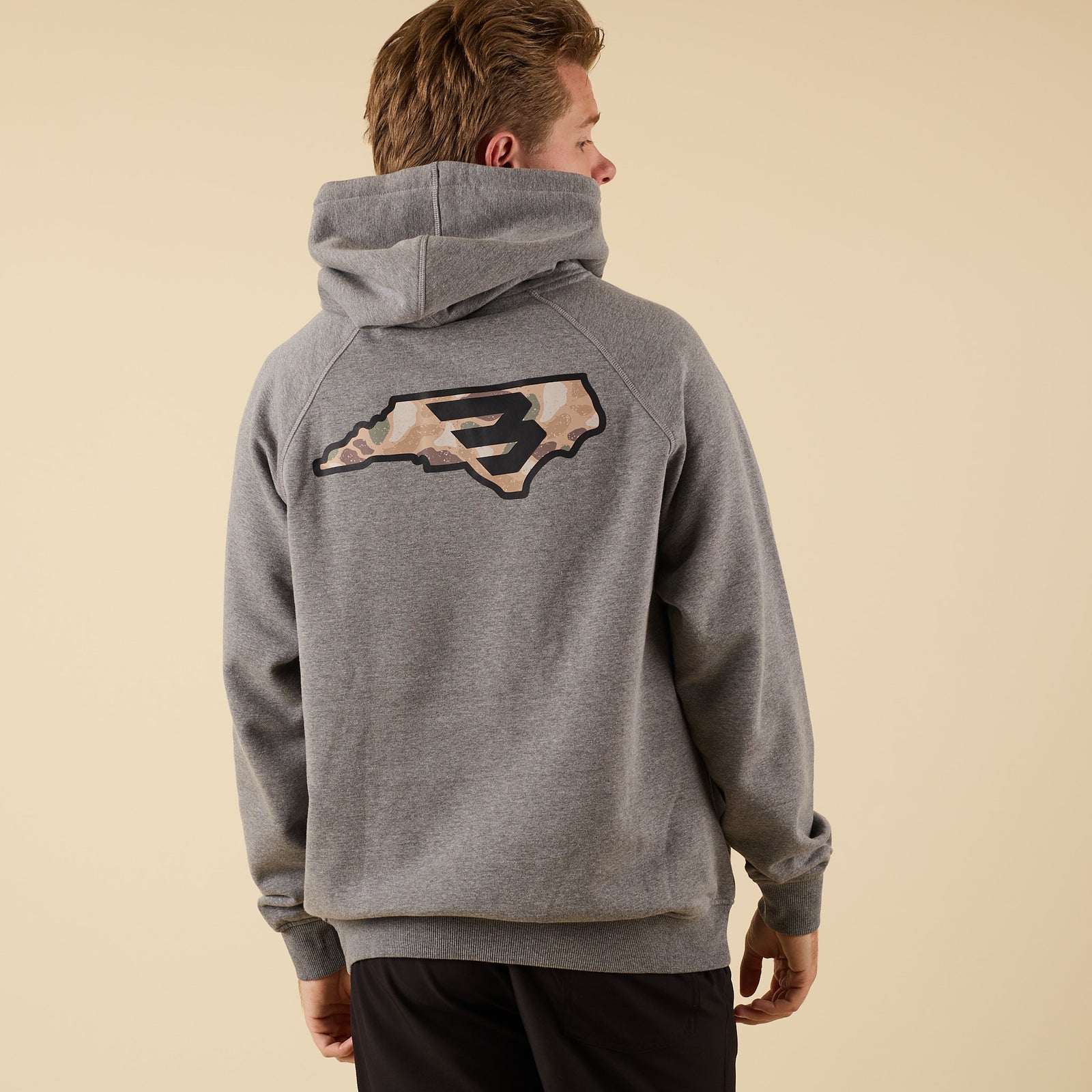 Rear angled view of a man wearing a gray hoodie with a large camo-pattern logo graphic centered between the shoulders.