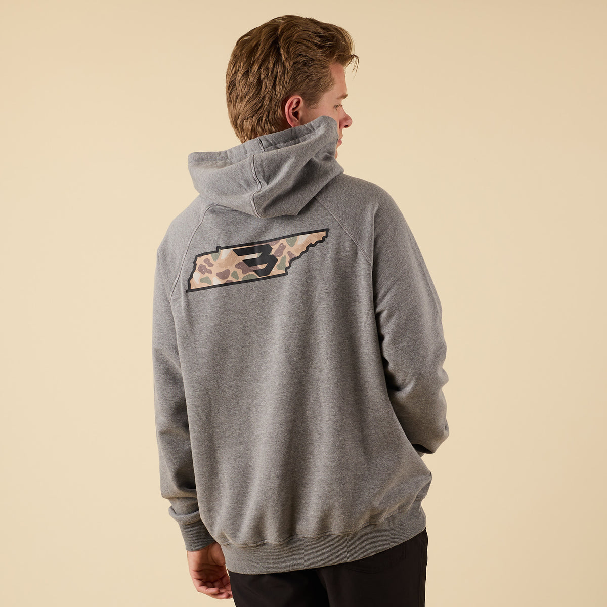 Rear angled view of a man in a gray Burlebo hoodie showing the large camo-pattern logo graphic on the back.