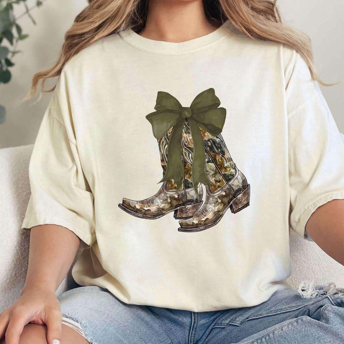 Ivory short sleeve t-shirt featuring graphic of camo print cowboy boots tied with an oversized olive green bow on the front center.