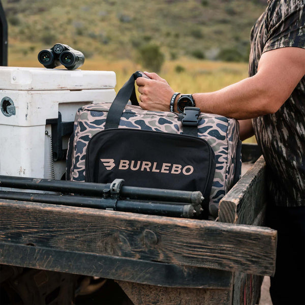 BURLEBO camo cooler bag in truck bed beside white cooler and binoculars, featuring black front pocket with logo and top carry handles.