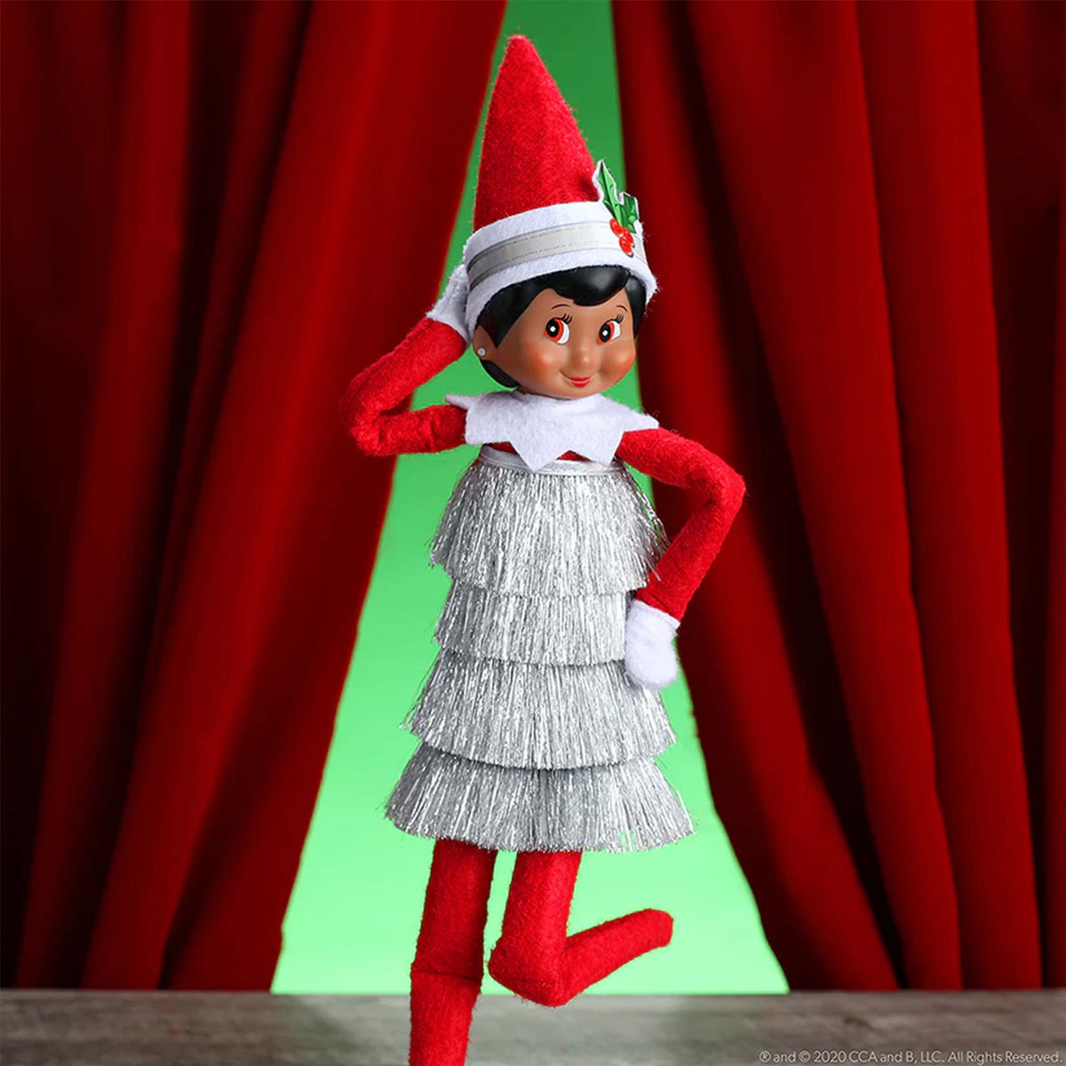 Elf on the Shelf with red hat and silver dress against a red curtain background