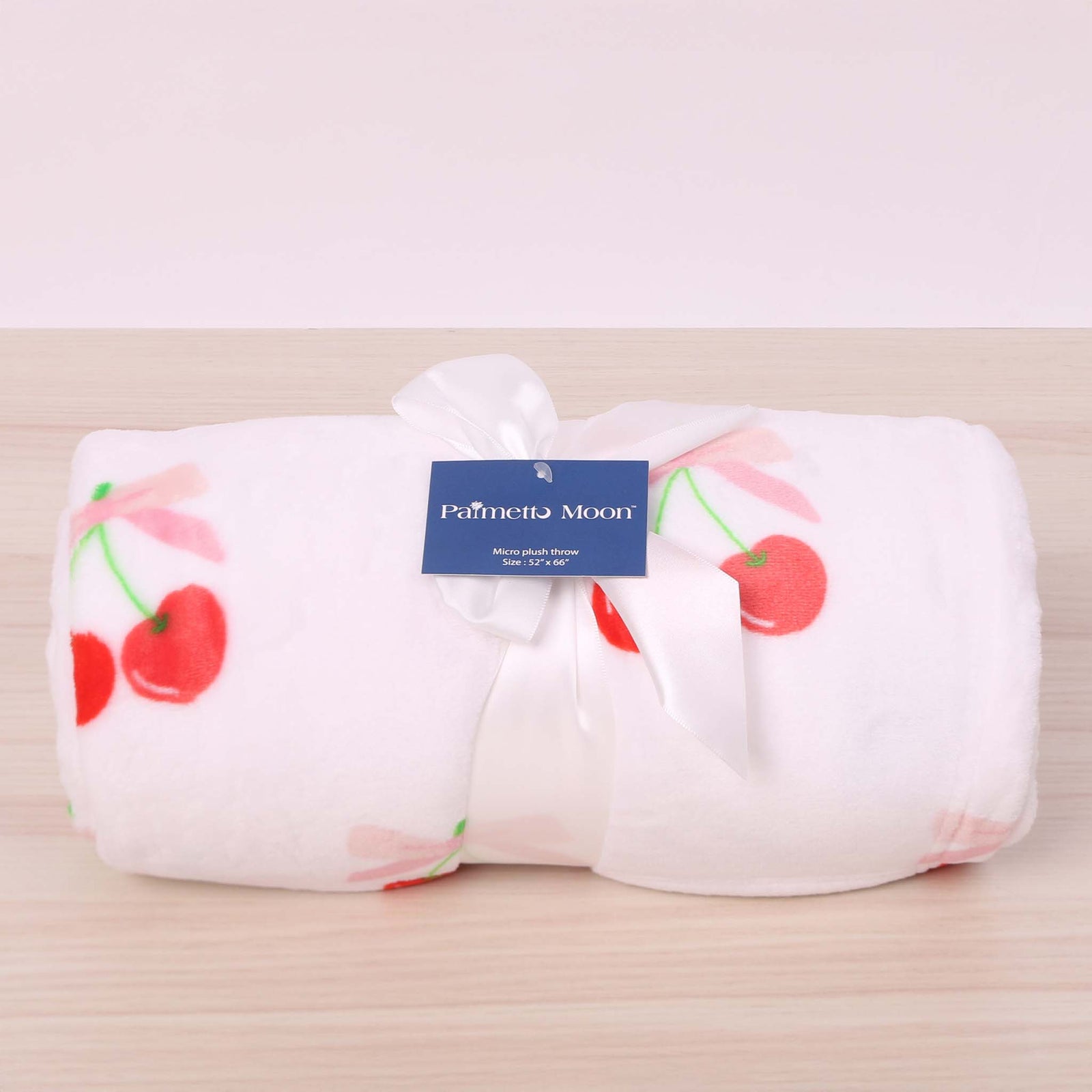 Folded white blanket with red cherry pattern and 'Palmetto Moon' label on a beige background