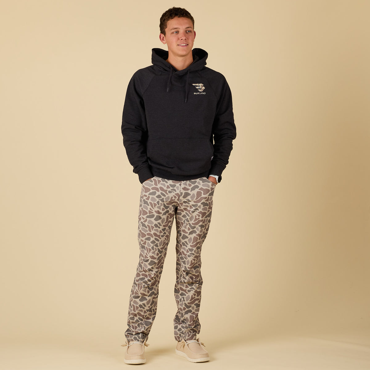 Full body view of a man wearing tan, brown, and gray deer camo pants styled with a black hoodie.