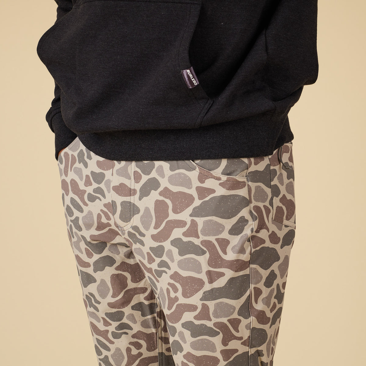 Close-up front view of men’s pants in tan, brown, and gray deer camo print with front pockets.