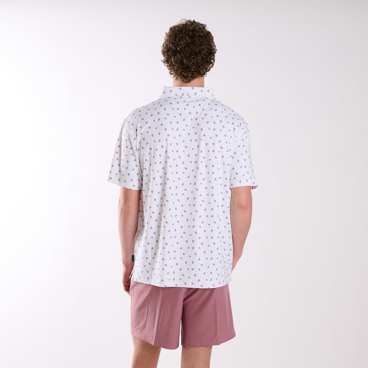 COAST Transfusion Icon Printed Polo in white with small transfusion cocktail icon print, shown from the back on a model wearing pink shorts against a light studio background.