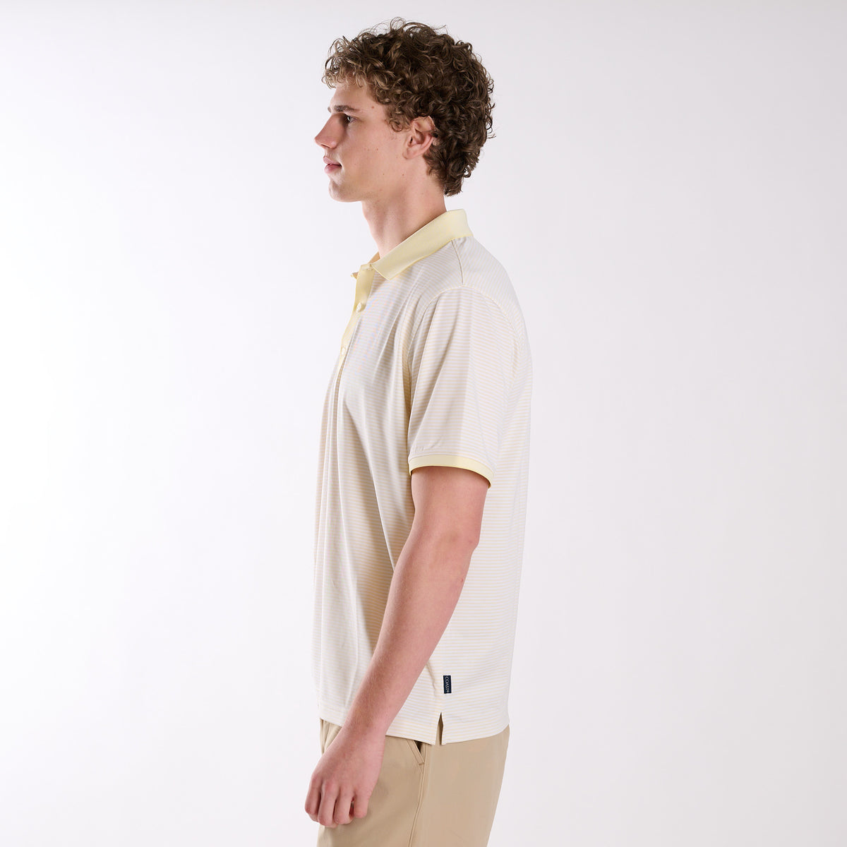 Side view of COAST micro-stripe performance polo in pale yellow with white stripes, worn by a model with khaki shorts against a neutral studio background.