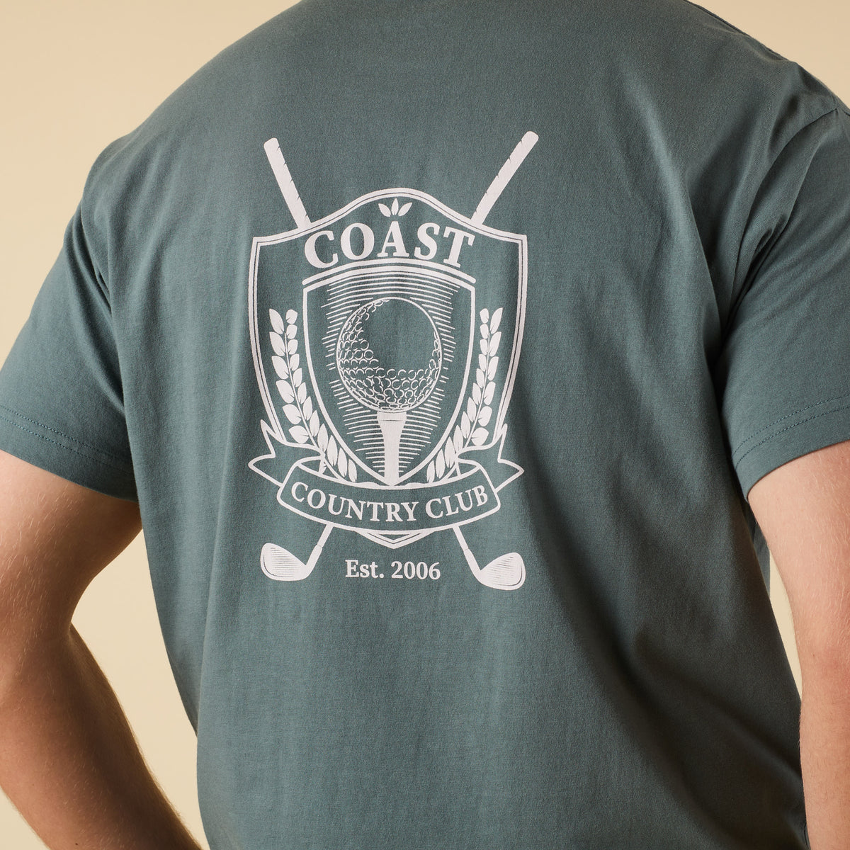 Close-up of COAST Cross Clubs Tee back crest design with golf ball on tee, crossed clubs, and "Country Club" banner.