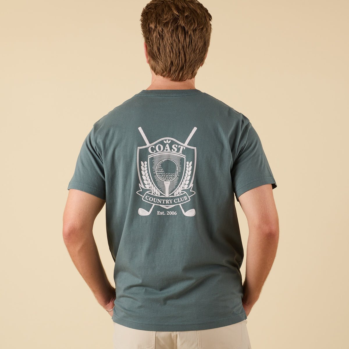 Back view of COAST Cross Clubs Tee in dark green with large white crest graphic featuring golf ball, crossed clubs, and laurel leaves.