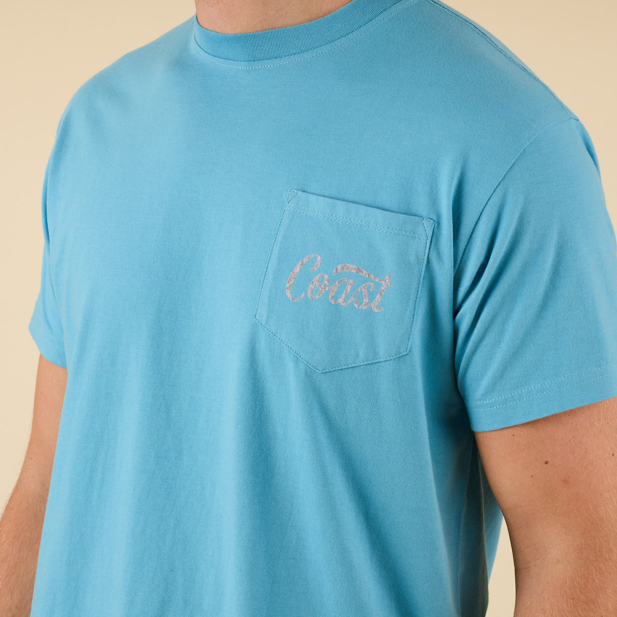 Close-up of COAST logo on front pocket of aqua blue Camo Label tee.