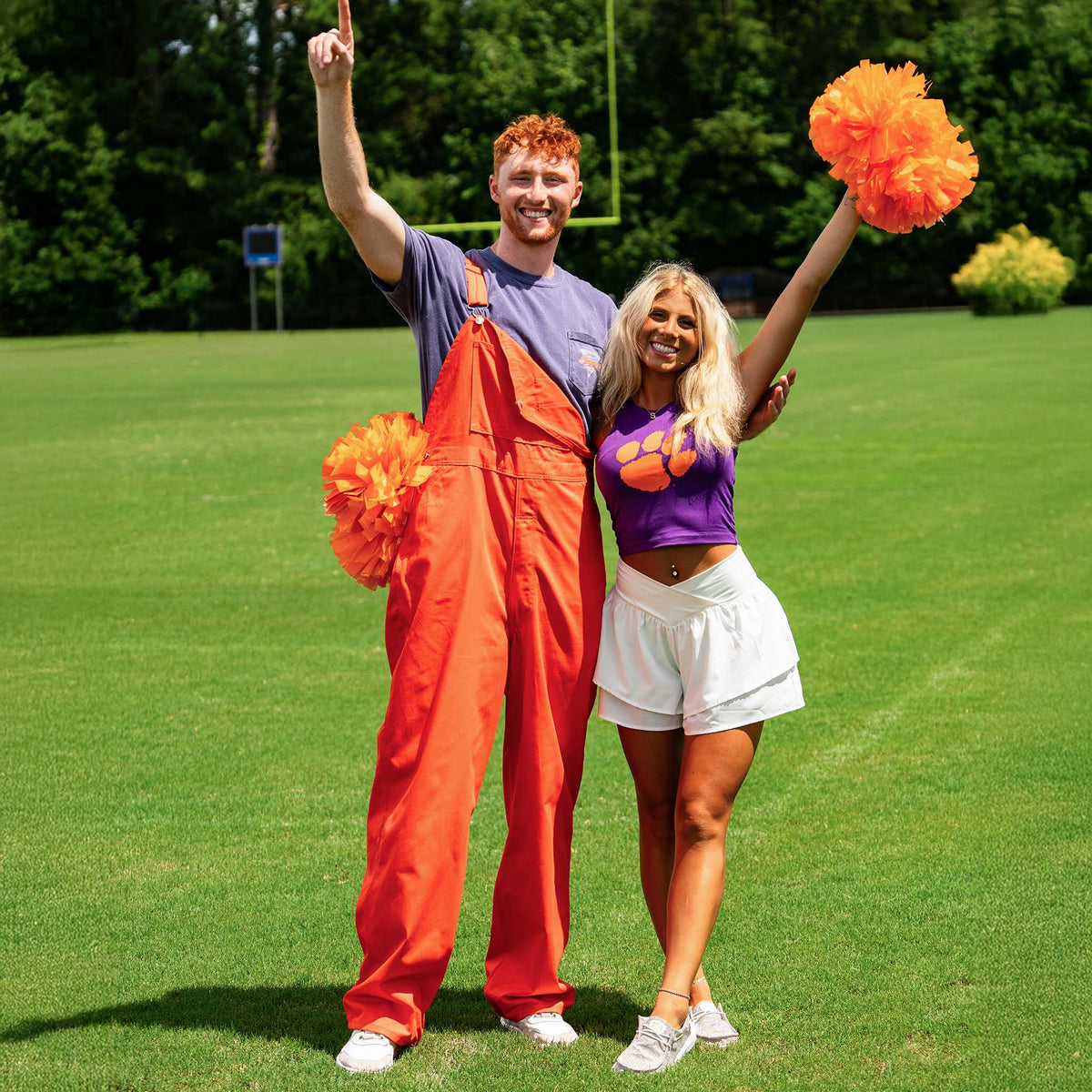 Man in bright orange overalls and woman in Clemson top and white skirt cheer on a football field, holding matching orange pom-poms and smiling.