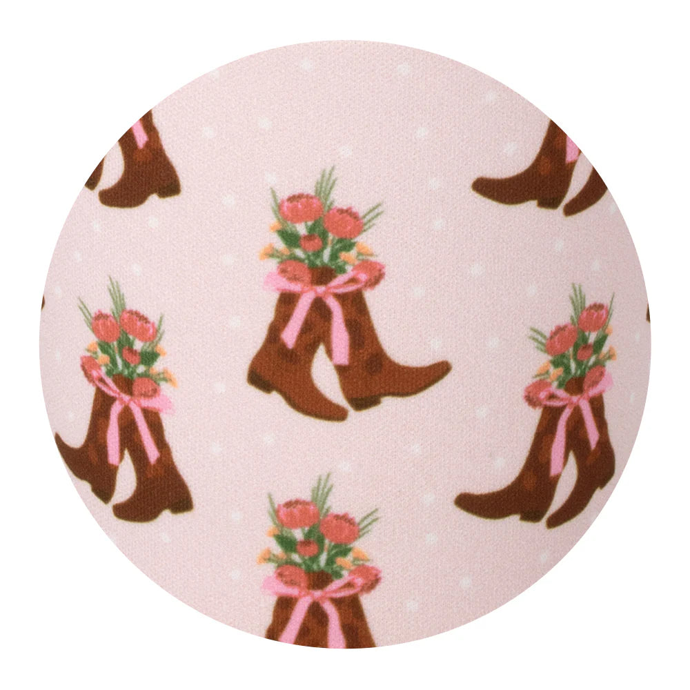 Close-up of Cowgirl Coquette print featuring brown cowboy boots filled with pink flowers and tied with pink ribbons on a light pink background with small white dots.