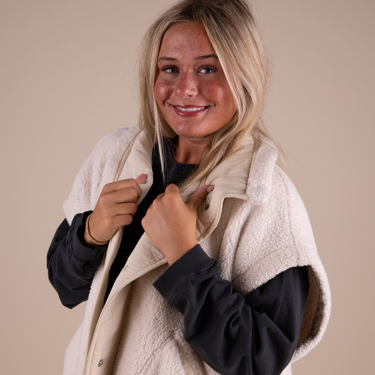 Woman wearing a dark long sleeve shirt under a beige sherpa vest.