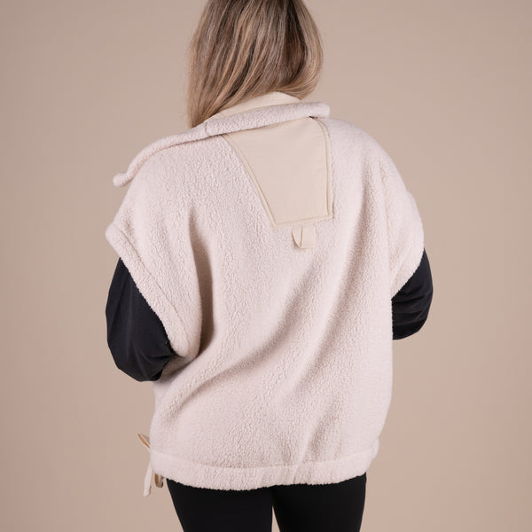 Woman wearing a dark long sleeve shirt under a beige sherpa vest.