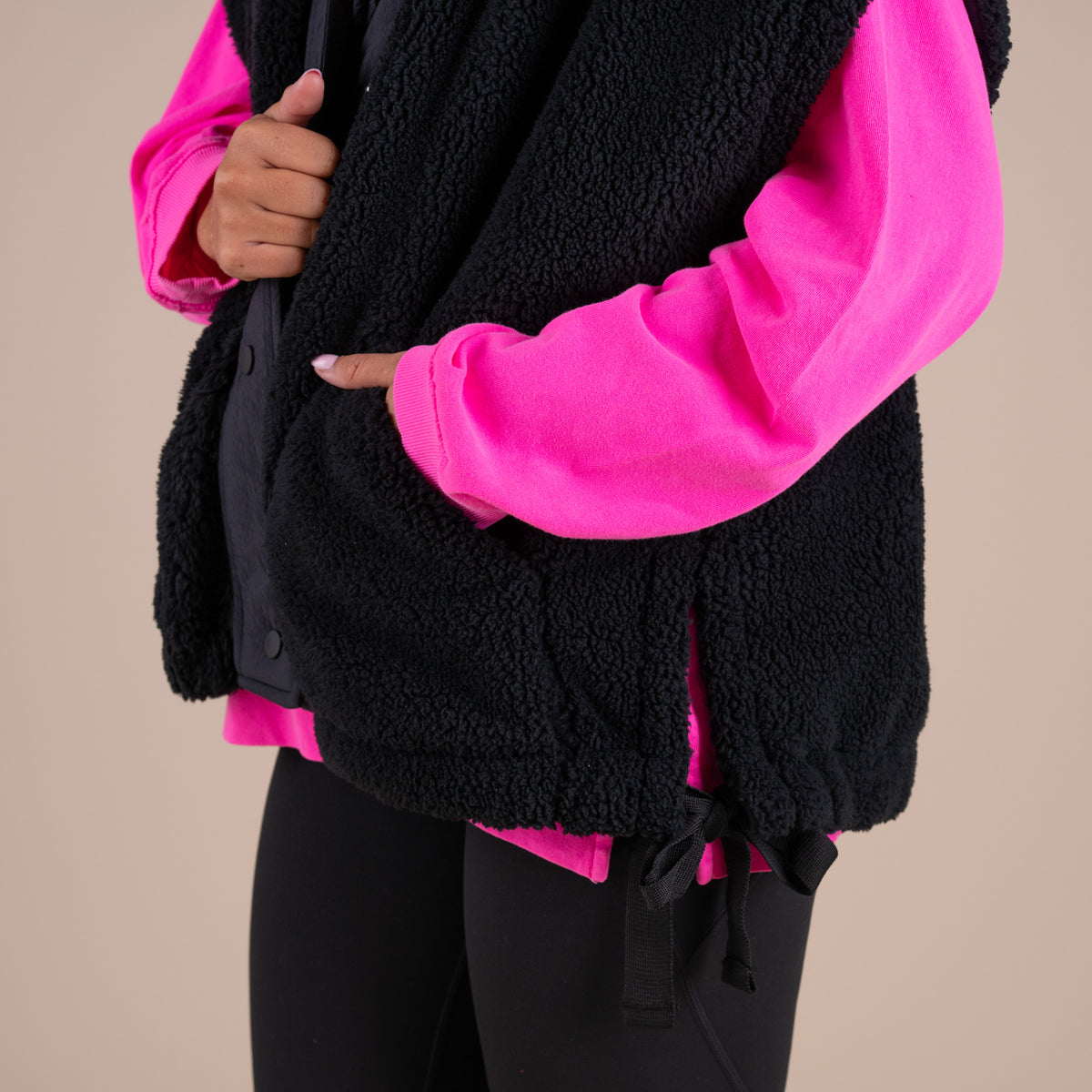 Person wearing a black fleece vest over a bright pink jacket on a beige background