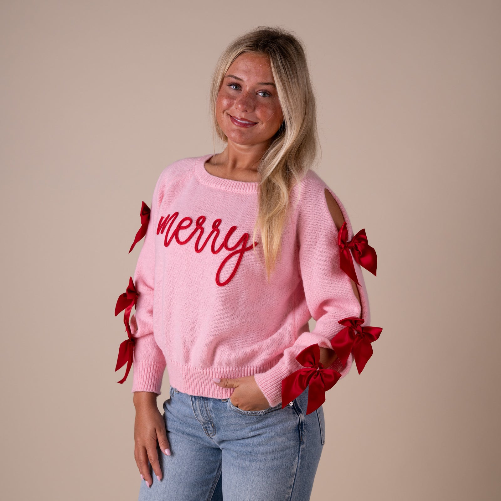 Woman wearing a pink sweater with red 'merry' text and bows, standing against a beige background