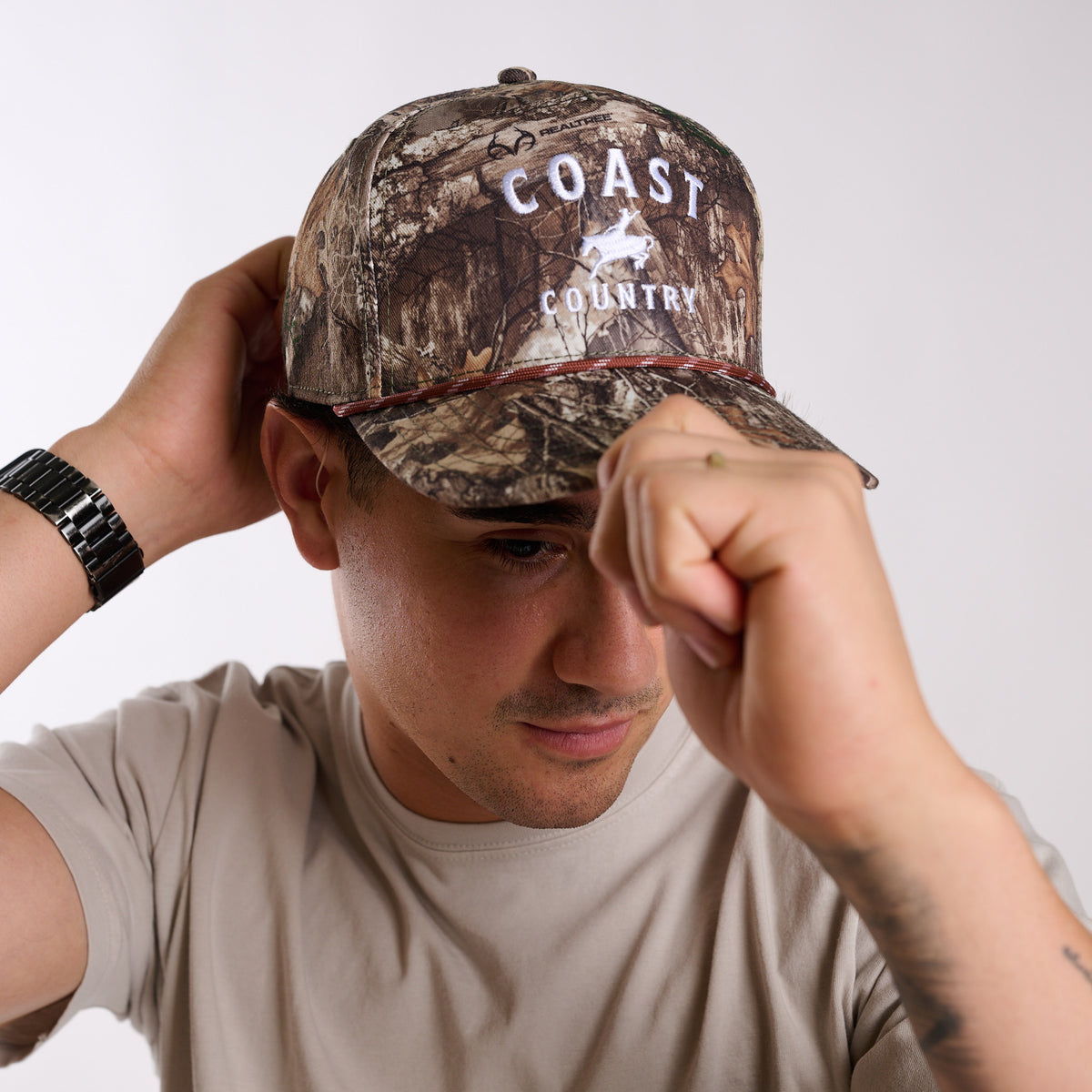 Person wearing a camouflage cap with 'Coast Country' logo on a plain background
