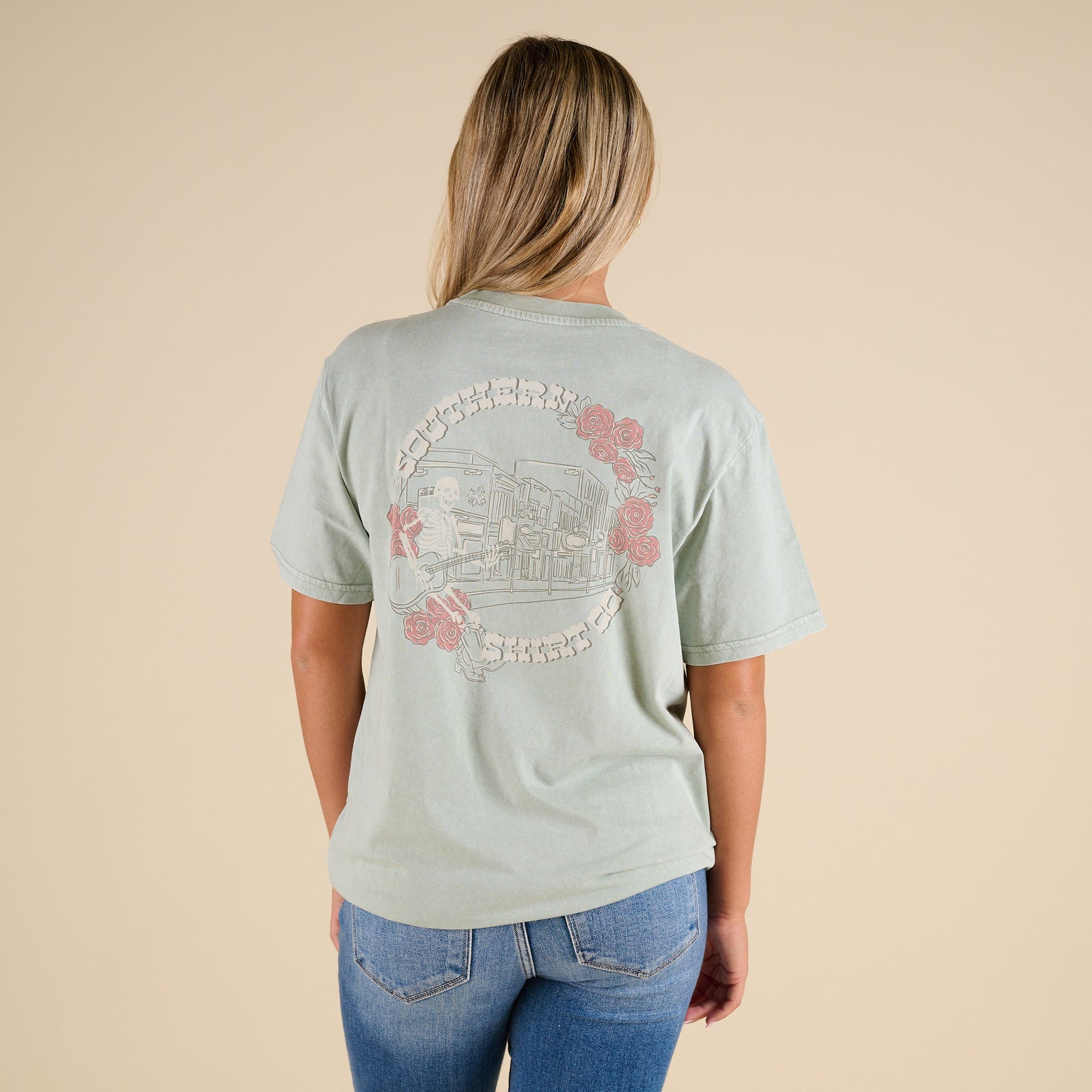 Person wearing a light green t-shirt with a graphic design on the back, standing against a beige background.