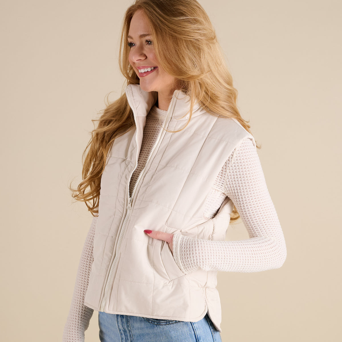 Woman wearing a beige vest over a white long-sleeve shirt on a beige background