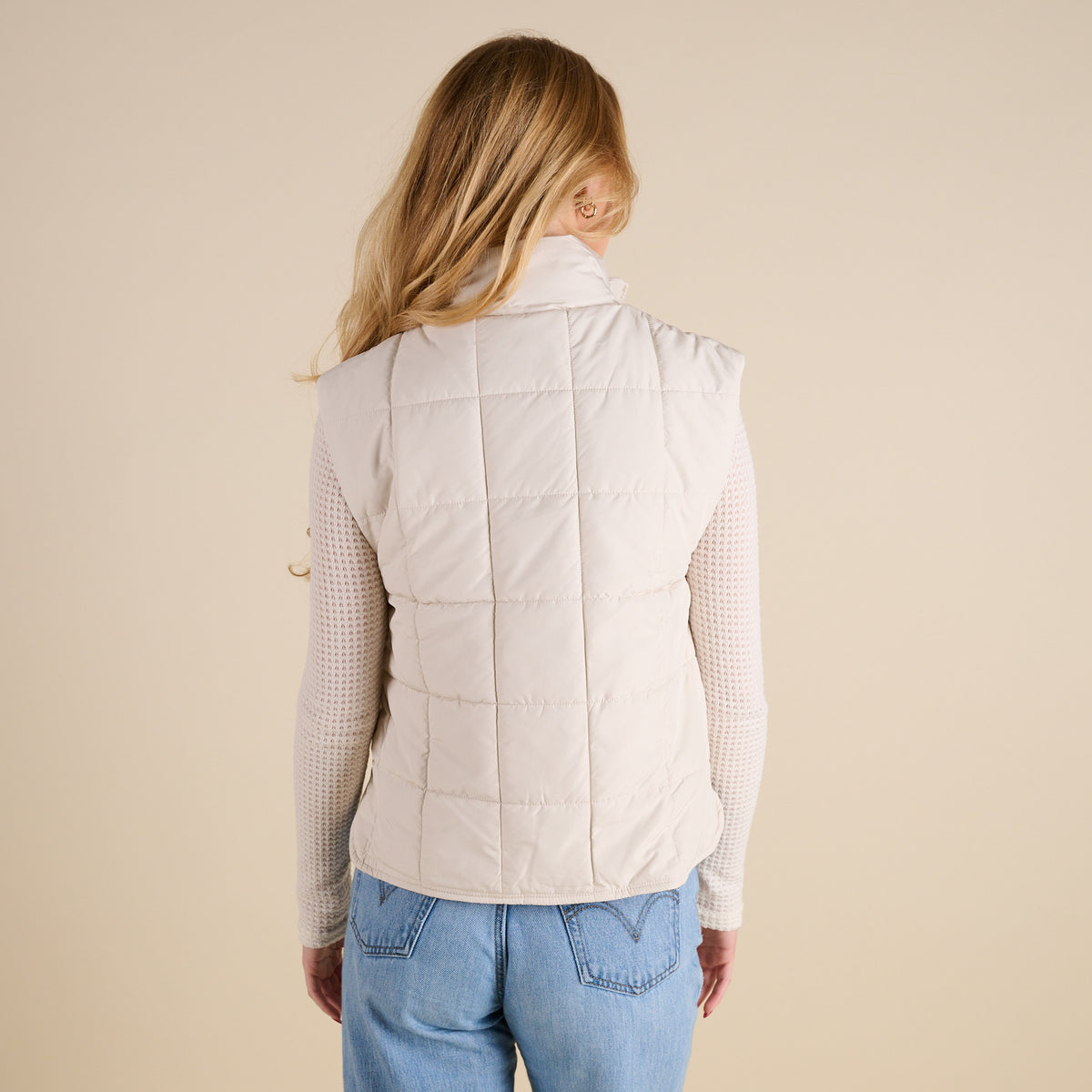 Person wearing a beige puffer vest and light blue jeans on a beige background