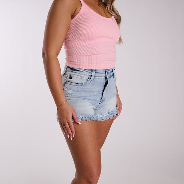 Woman wearing light wash high rise frayed hem denim shorts with a pink top on a white background