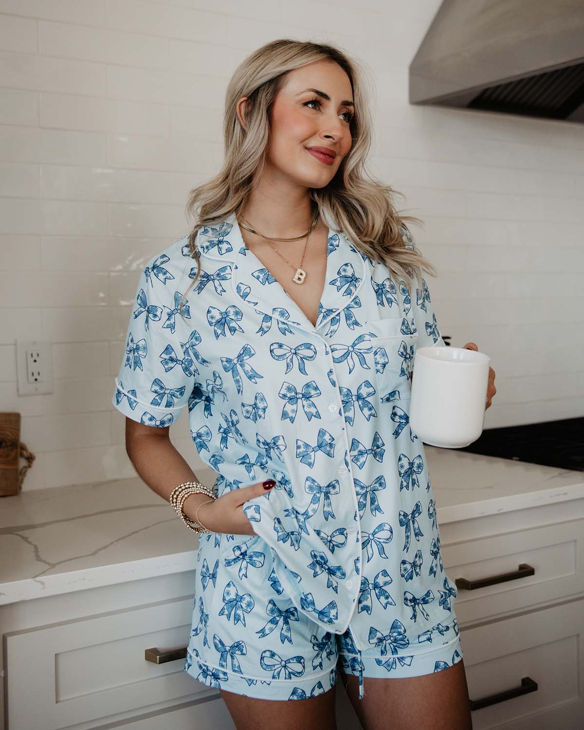Woman in a blue bow-patterned outfit holding a white mug in a kitchen.