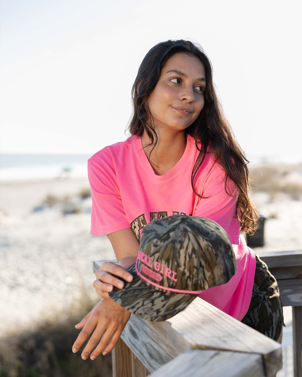 Local Girl camo rope hat with pink logo held by woman wearing bright pink t-shirt on beach boardwalk.