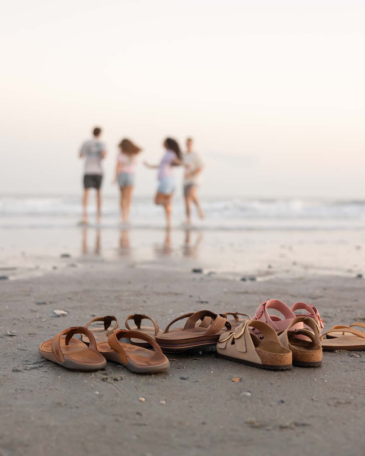 Assorted flip flop sandals arranged on sandy beach with group of people walking in ocean in background.