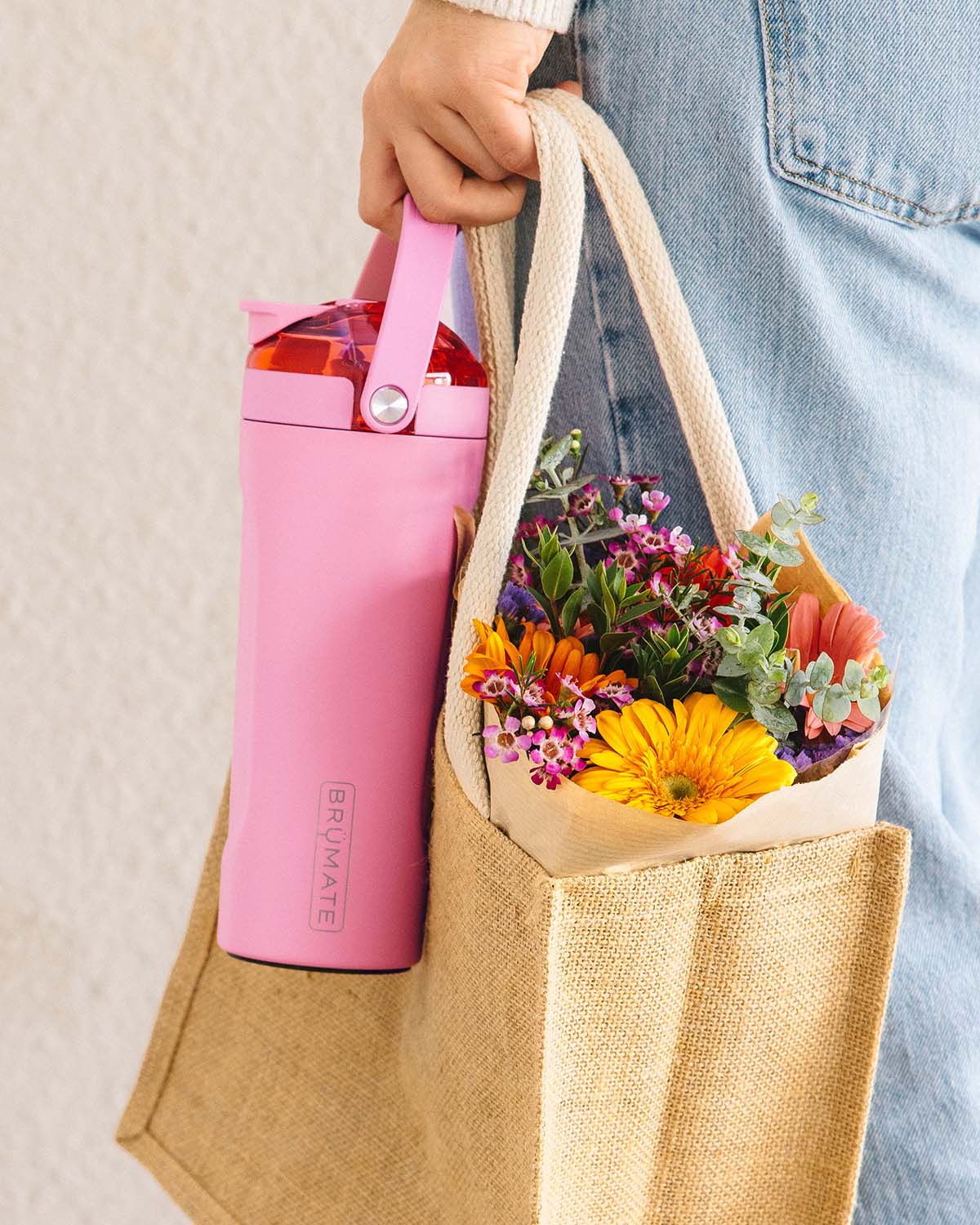 Pink BrüMate insulated bottle with handle carried in hand alongside a jute tote filled with colorful flowers.
