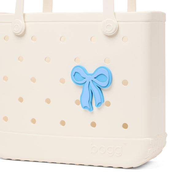 Bogg Bag Bogg Bit blue bow charm attached to cream Bogg Bag with perforated holes.
