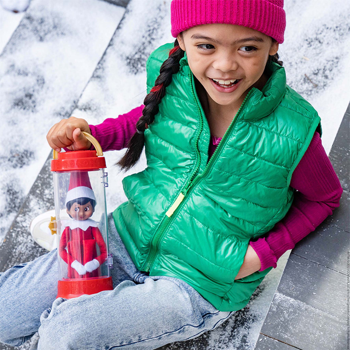 Child in green vest and pink hat holding a container with an elf on the shelf toy outdoors.
