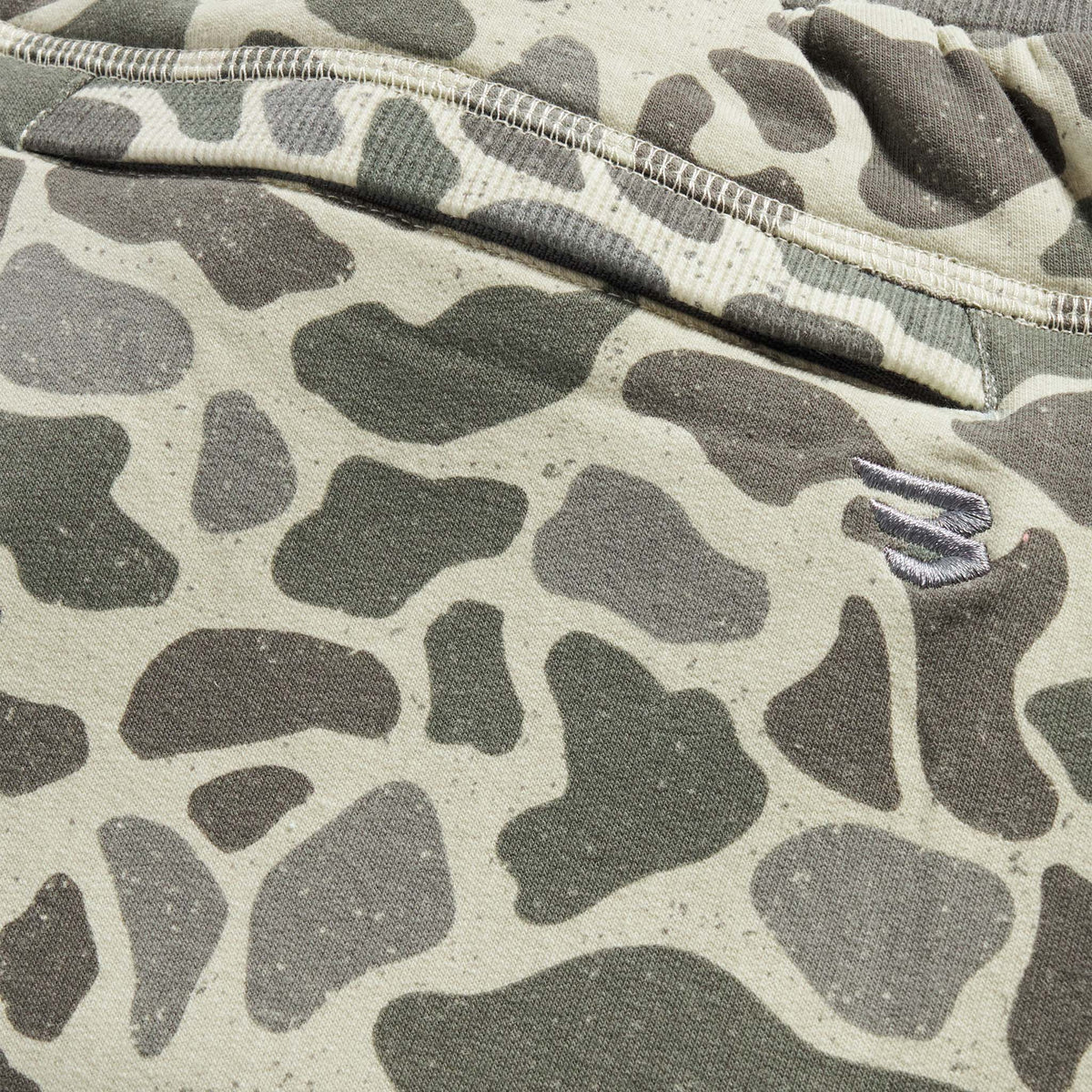 Close-up view of camo joggers featuring green and taupe abstract camo print with embroidered logo detail near the seam.