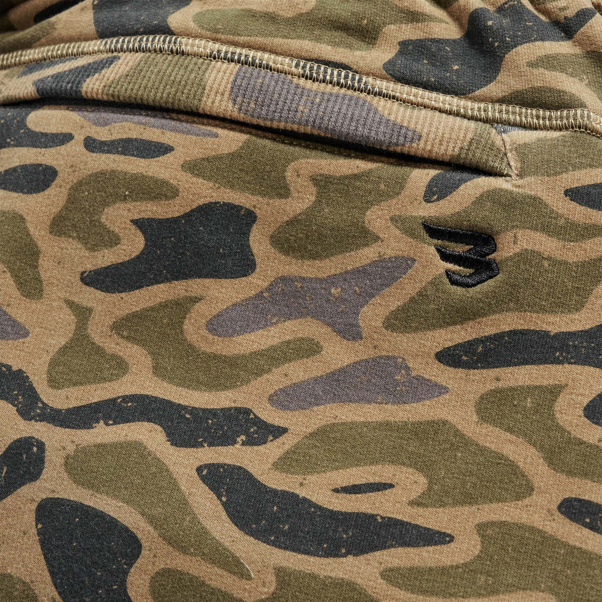 Close-up of men’s camo jogger pants in green, tan, and gray with tonal embroidered logo detail above the pocket.