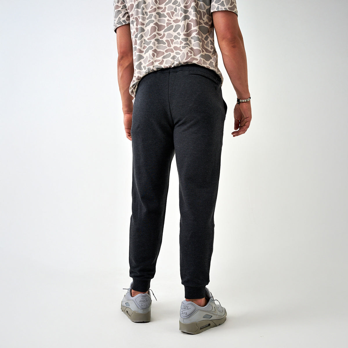 Back view of men’s dark charcoal joggers with elastic waistband and ribbed ankle cuffs, paired with light gray sneakers.