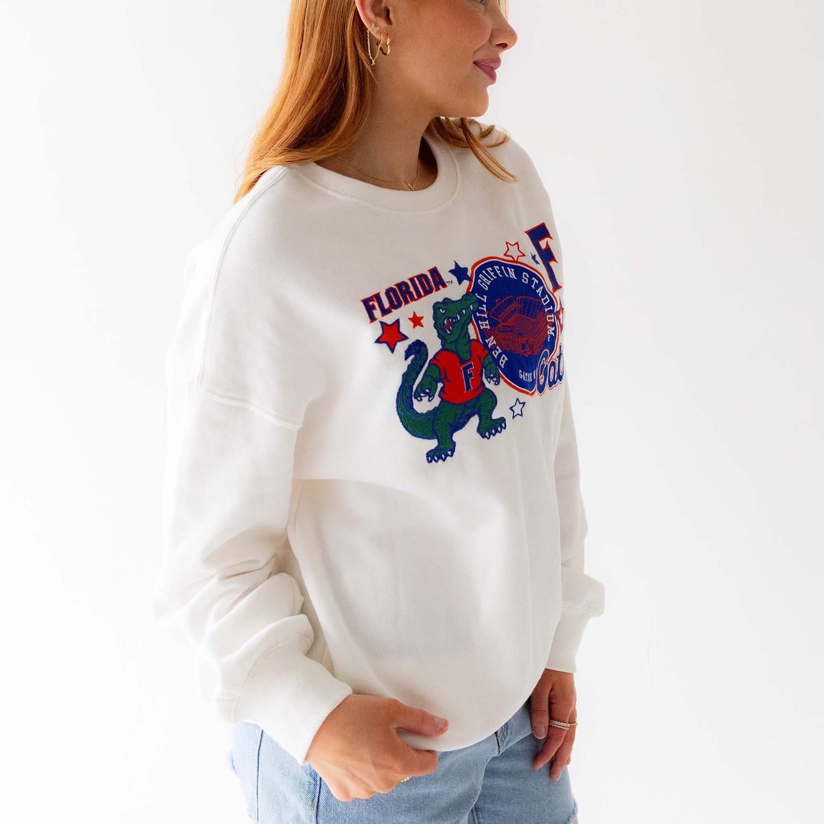 Side view of woman wearing white Florida Gators crewneck sweatshirt with colorful embroidered mascot, stadium patch, and stars.
